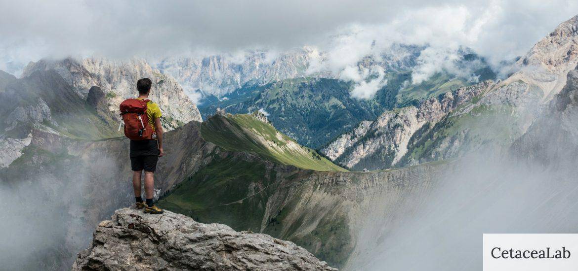 man with red backpack standing on cliff facing mountains under white sky during daytime