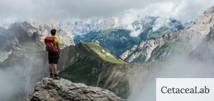 man with red backpack standing on cliff facing mountains under white sky during daytime