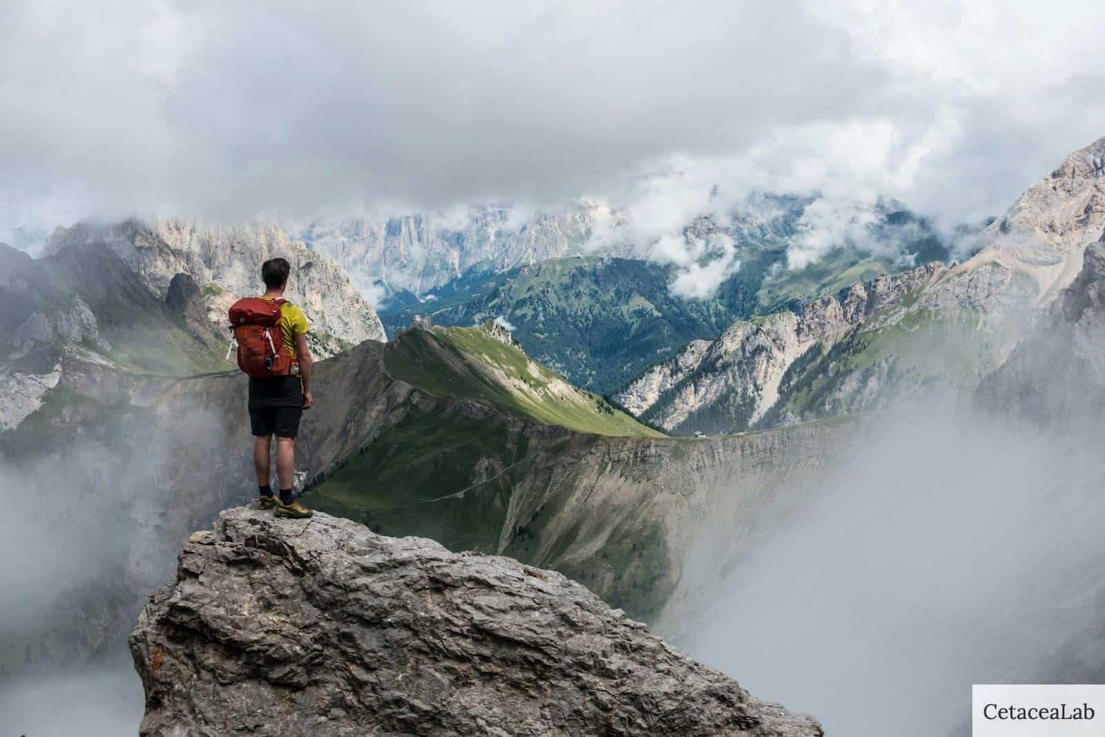 man with red backpack standing on cliff facing mountains under white sky during daytime